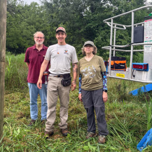 Two men and one woman standing in a grassy area next to a steel safety bucket with equipment on it.
