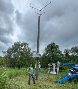 A man standing next to a very tall radio receiving tower with four antennas at the very top